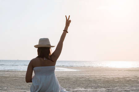 Back side of happy woman sitting on tropical sea and beach showing two fingers or victory sign during sunset. Young woman enjoy life vacation and relaxing in paradise island in vintage tone.の写真素材
