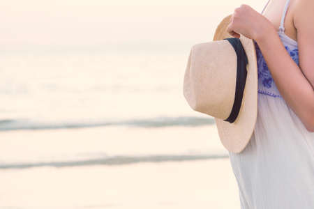Cropping image of attractive and happy woman standing on tropical beach and hand holding straw hat in summer vacation with copy space. Young girl wear white dress walking alone at sea in sunset.の写真素材