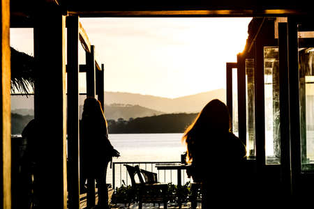 Silhouette of woman walking into cafe side the sea with beautiful blazing sunset landscape at mountain, sea and orange sky background with awesome sun golden reflection. Sunset view at restaurant.の写真素材
