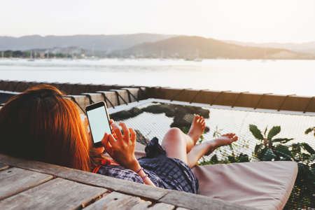 Young woman using black smartphone with blank desktop screen while lying side the sea with evening sunset background. Mockup image of woman hand holding phone chatting online message with friends.の写真素材