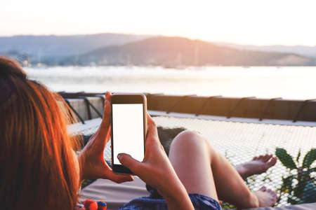 Young woman using black smartphone with blank desktop screen while lying side the sea with evening sunset background. Mockup image of woman hand holding phone chatting online message with friends.の写真素材