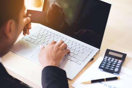 Businessman hand holding credit card and using laptop on desk with document and calculator. Man hand use card for online shopping and online payment cashless society concept.の写真素材