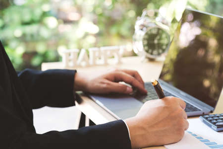Close up businessman hand holding pen and taking note in paper and laptop on wood desk in workplace. Success business man checking report profit on paper with rising charts, discussing company growth.の写真素材