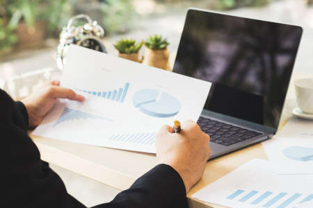Businessman hand holding business documents of graph financial diagram on wooden desk with laptop in office. Success business man working and checking report profit on paper with rising charts.の写真素材