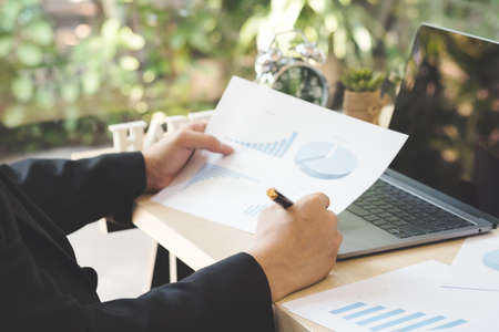 Young businessman holding business documents of graph financial diagram on wooden desk with laptop in office. Success entrepreneur working and analyze report profit on paper with rising charts.の写真素材