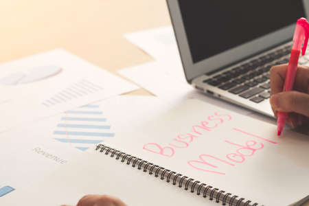 Young businessman hand writing word Business Model on notebook with pink marker pen, laptop and business document on wooden table.の写真素材