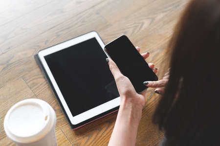 Close up view on hand of young woman holding smartphone with blank screen and digital tablet pc and white coffee cup on wooden table in cafe. Business woman using devices at home for working.の写真素材