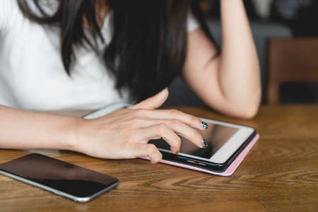 Close up image of young asian woman in casual using digital tablet and smartphone on wooden table in cafe. Business woman reading message and social media on tablet. Device and technology concepts.の写真素材