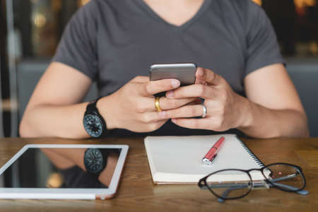 Cropped image of young man in casual using smartphone, digital tablet and notebook on wooden table at cafe. Student college male texting message on mobile phone. Lifestyle and education concepts.の写真素材