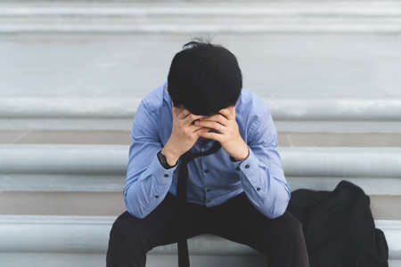 Young asian businessman sitting on concrete stairs and holding head with hand are stressed with work and life issues. He thinks to commit suicide when he know business is failing and unemployed.の写真素材