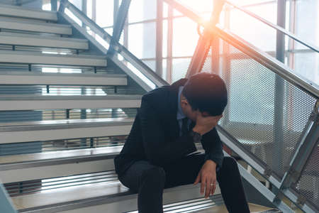 Young asian businessman sitting on stairs are stressing when he know business is failing and unemployed in office building. Depressed emotional after receiving bad news. Dismissed or deposed employee.の写真素材