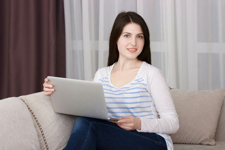 Happy young woman is relaxing on comfortable couch and using laptop at homeの写真素材