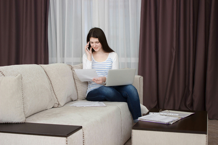 Happy young woman is relaxing on comfortable couch and using laptop at homeの写真素材