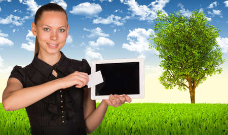 Beautiful businesswoman holding blank tablet PC and blank business card in front of PC screen. Green meadow, sky and tree as backdropの写真素材
