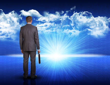 Businessman with briefcase standing against digitally generated spacy blue landscape with rising sun and cloudy skyの写真素材