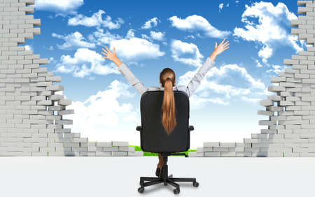 Businesswoman sitting on the office chair with her hands outstretched, in front of broken wall with blus sky and green field behind it.の写真素材