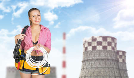 Woman in tool belt, holding coils of cable, looking, at camera, smiling. Sky, clouds and power plant...