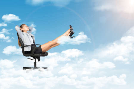 Businesswoman leaning back in office chair standing on cloud, with her feet up on smaller cloud, looking at light shining from aboveの写真素材