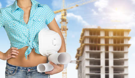 Woman in shirt and shorts holding white helmet and rolls of paper. Cropped image. Building under construction in backgroundの写真素材
