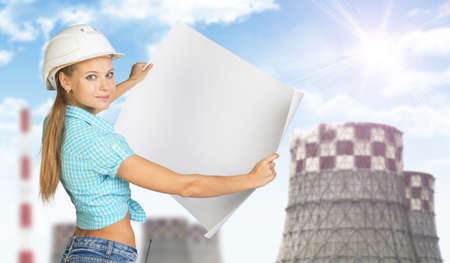 Woman in white helmet holding white paper sheet. Looks at camera, smiling. Chimneys as backdropの写真素材