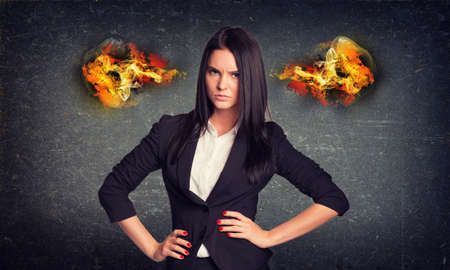 Angry businesswoman standing with arms akimbo, fire from ears. Concrete wall in backgroundの写真素材