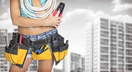 Woman in tool belt with different tools holding cable and pliers. Cropped image. Buildings and gray...