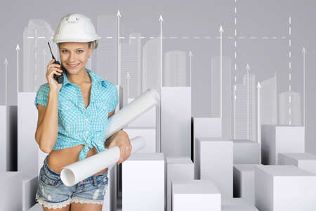 Beautiful girl in working clothes holding walkie-talkie and paper scrolls, looking at camera, smiling. Minimalistic city of white cubes with arrows on gray backgroundの写真素材