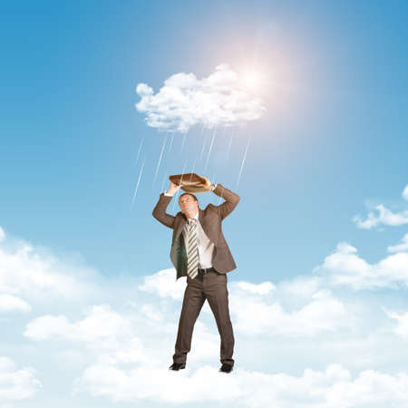 Businessman in suit standing on cloud and covered using briefcase from rain. Sky with sun as backdropの写真素材