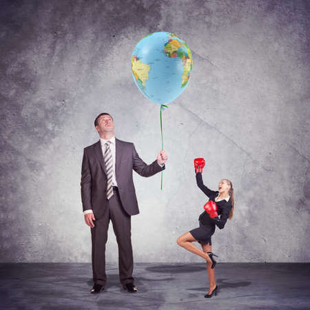 Conceptual Businessman Holding and Looking at the Balloon, with World Map Print, with a Small Businesswoman Posing in Boxing Gloves on the Side. Captured in Studio with Weathered Wall Backgroundの写真素材