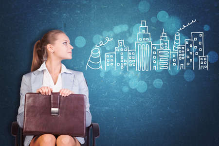 Young Businesswoman Sitting in Chair with Briefcase in Lap, Looking to the Side at Illustration of City Skyline in Aspirational Concept Imageの写真素材