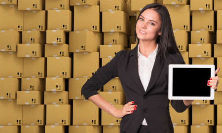 Smiling young woman holging tablet and looking at camera on abstract cardboard boxes set  backgroundの写真素材