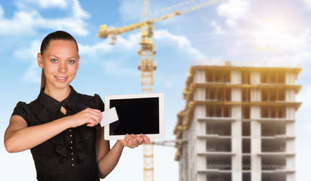 Young woman holging tablet and blank card and looking at camera. Construction with building crane on backgroundの写真素材