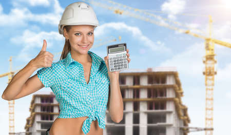 Smiling young woman in hard hat holding calculator and looking at camera, close-up. Industrial backgroundの写真素材