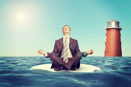 Businessman sitting in lotus position on small island in sea and looking upの写真素材