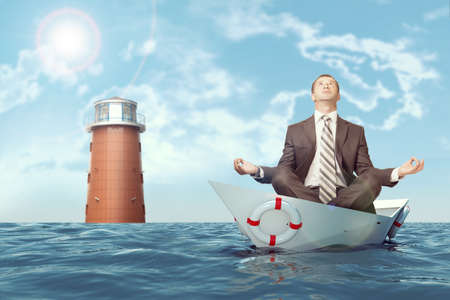 Businessman sitting in lotus position in paper boat and lighthouse in sea and looking upの写真素材