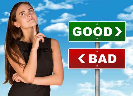 Thoughtful businesswoman standing near colorful good and bad signboard and looking up on blue sky backgroundの写真素材