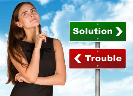 Thoughtful businesswoman standing near colorful solution and trouble signboard and looking up on...