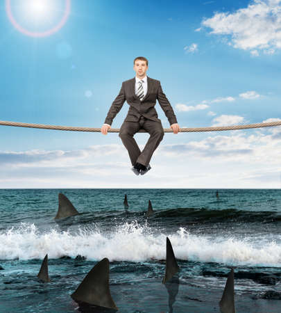 Businessman sitting on rope above ocean with sharks and looking at cameraの写真素材