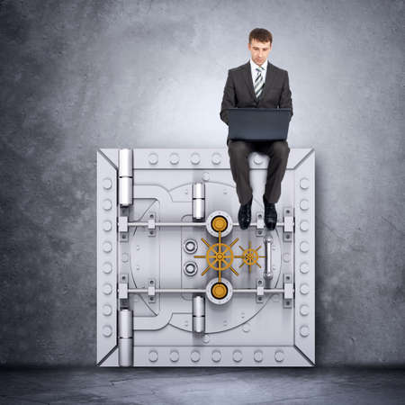Businessman working on laptop and sitting on safe door, front view