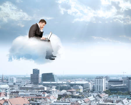 Sad businessman working on laptop and sitting on cloud above cityの写真素材
