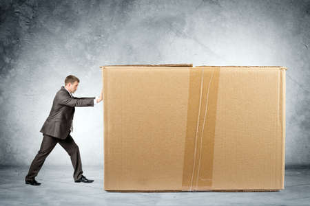 Businessman pushing big brown carton box on grey wall background