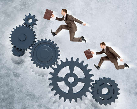 Two businessmen running on wheel gears on grey wall backgroundの写真素材