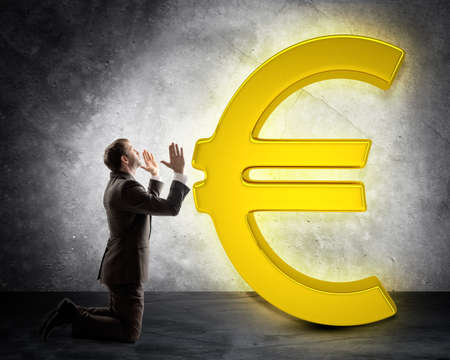 Businessman on knees in front of euro sign on grey wall backgroundの写真素材