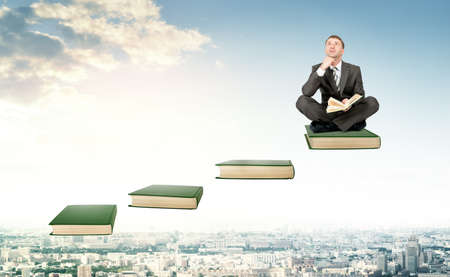 Young man in suit sitting on ladder steps with book in hands above cityの写真素材