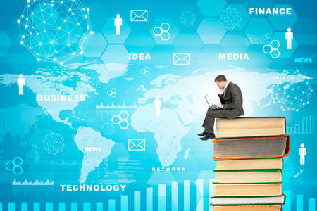 Businessman with laptop sitting on stack of books on blue background with world mapの写真素材