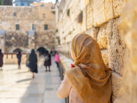 A woman draped in a hijab leans thoughtful against a textured stone wall, observing passersby.の素材