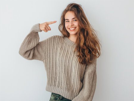A smiling young woman in a cozy sweater confidently points at herself against a white backdrop.の素材