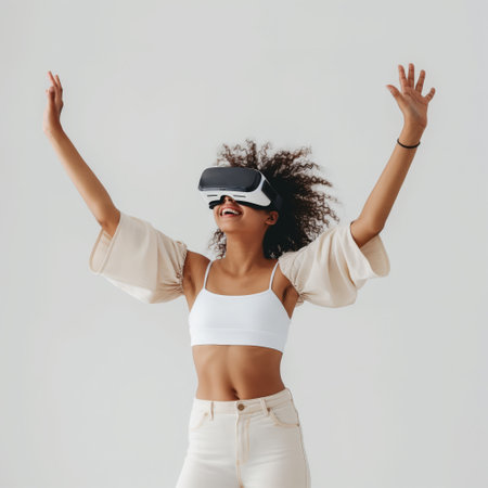 A woman with curly hair smiling and raising hands while wearing virtual reality goggles against a neutral background.の素材