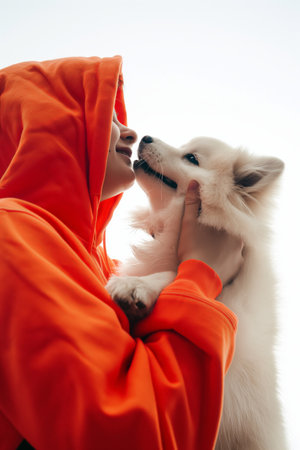 A person in an orange hoodie is affectionately touching noses with a white fluffy dog against a clear backdrop.の素材