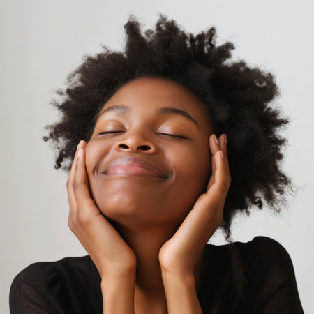 Close-up of a content young woman with her eyes closed, hands gently cradling her face, and a serene smile.の素材